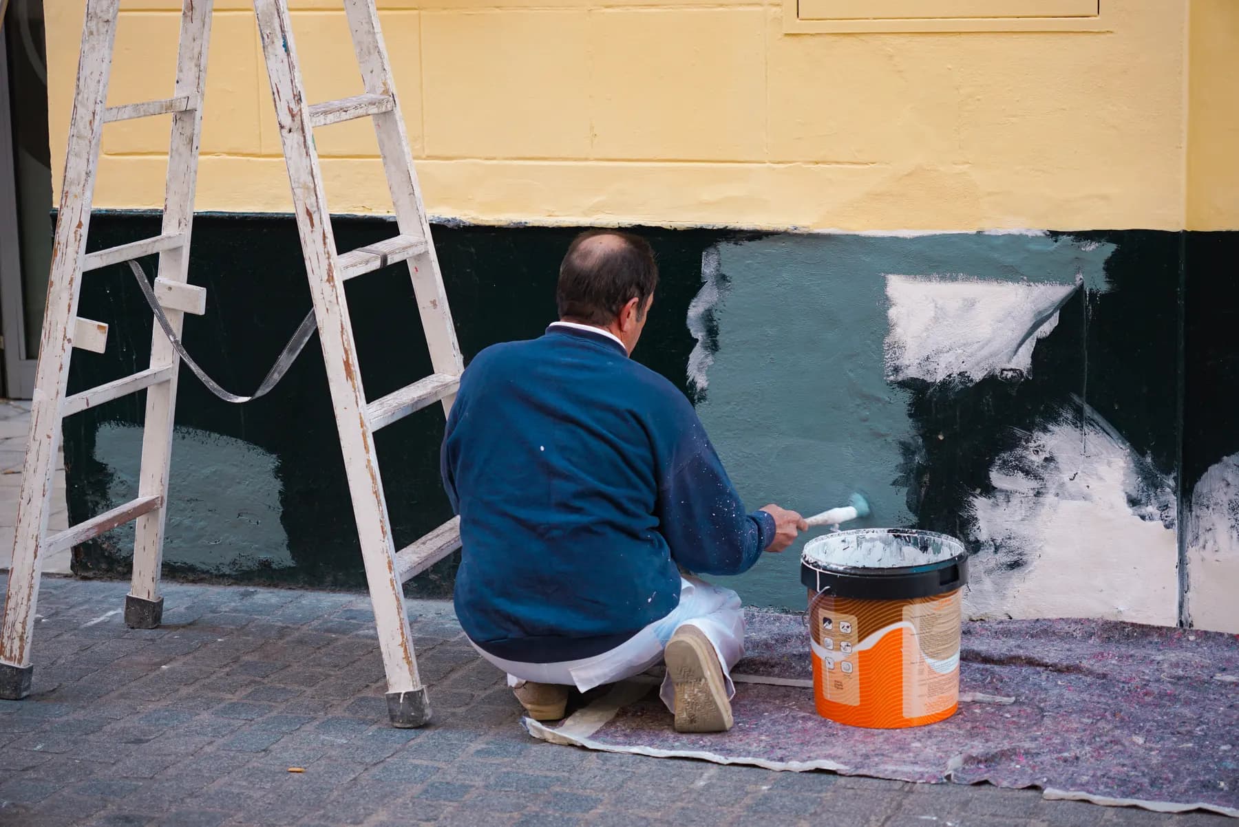 Painter kneeling beside a ladder rolling fresh color onto an exterior wall